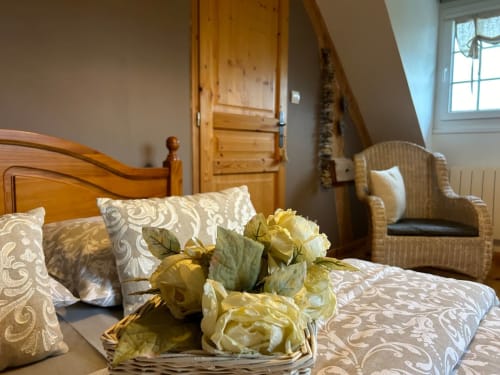 Chambre raffinée au Clos Mahiel avec lit en bois, coussins à motifs, fleurs et fauteuil en osier près de la fenêtre