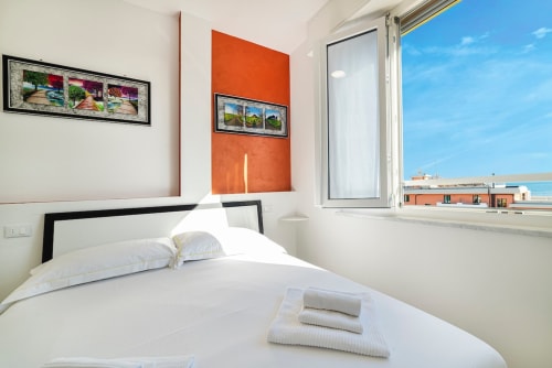 Modernes Schlafzimmer in der Residenza la Pietra mit Meerblick, weißem Bett, orangefarbener Akzentwand und sichtbarer Küstenlandschaft