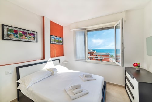 Modernes Schlafzimmer in Pietra Ligure mit Meerblick, weißem Bett, orange akzentuierter Wand und Balkon mit Blick auf das Mittelmeer