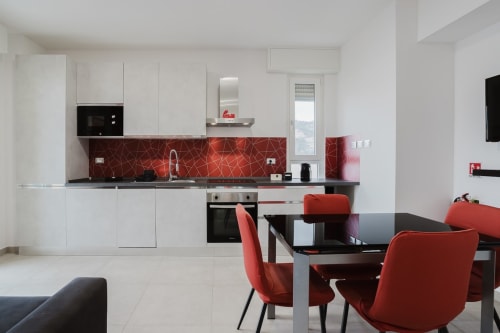 Moderne Küchen-Esszimmer mit roten geometrischen Fliesenmustern, weißen Schränken und roten Stühlen am schwarzen Tisch in Pietra Rossa