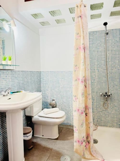 Baño moderno en Apartamentos Rocamar con paredes de azulejos mosaico, lavamanos pedestal, inodoro y ducha abierta con cortina floral
