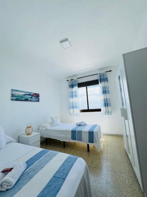 Dormitorio luminoso en Apartamentos Rocamar con dos camas, ropa de cama con rayas azules, ventana con cortinas y cuadros de océano.