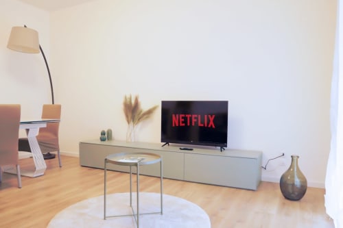 Modern living room with TV showing Netflix, light wood flooring, gray media console, minimalist decor at Treviso Stays