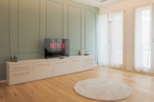 Modern living room with TV showing Netflix, light wood furniture, pale green paneled walls, wooden floors, and glass doors