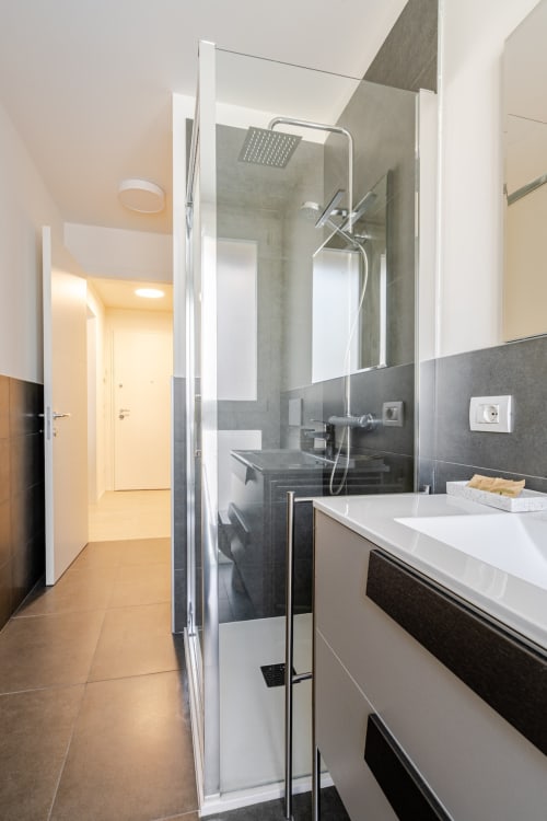 Modern bathroom in Treviso apartment with glass shower enclosure, white sink vanity, and dark tile accents