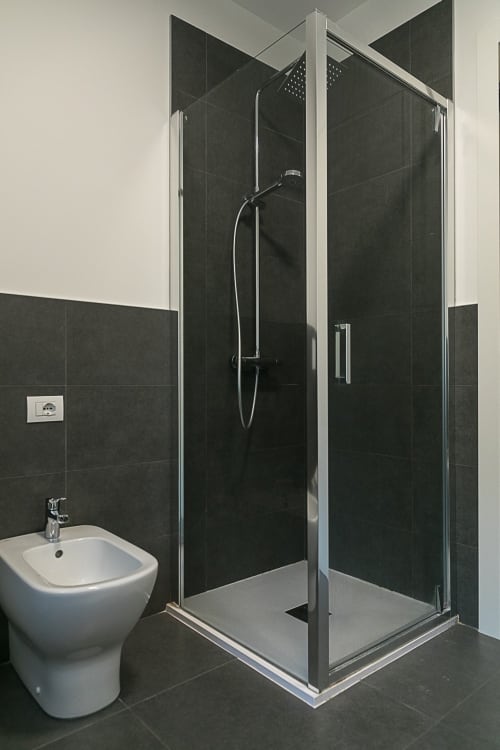 Bagno moderno con piastrelle nere, box doccia in vetro, soffione a pioggia, wc e bidet bianchi a Treviso