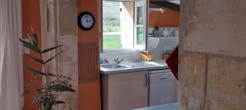 Bright cottage kitchen at La Barrat with orange walls, sink, dishwasher, clock, and garden view through glass doors