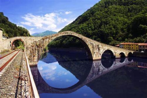 Historic stone bridge spanning calm river near Lucca, Italy with lush green mountains and railway tracks