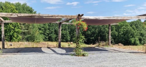 Pergola rustique en bois avec fleurs suspendues au-dessus d'une terrasse en gravier à la Domaine de la Grande Décision, entourée de verdure luxuriante