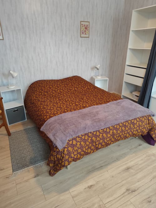 Chambre douillette de l'appartement Accolade à Châteaudun avec couvre-lit motifs, table de nuit blanche et parquet.