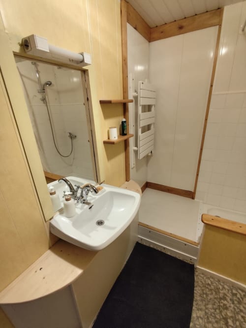 Salle de bain moderne avec lavabo blanc, cabine de douche, étagères en bois et sèche-serviettes chauffant à l'appartement Accolade à Châteaudun