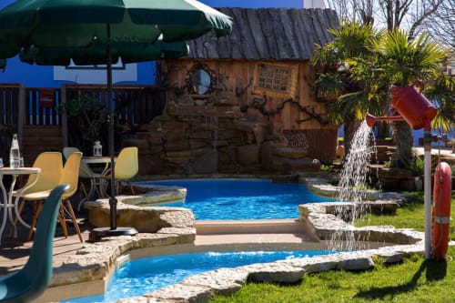 Ninho Saloio's tropical-themed pool area with blue water features, wooden cabin, palm trees, and shaded seating in Venda do