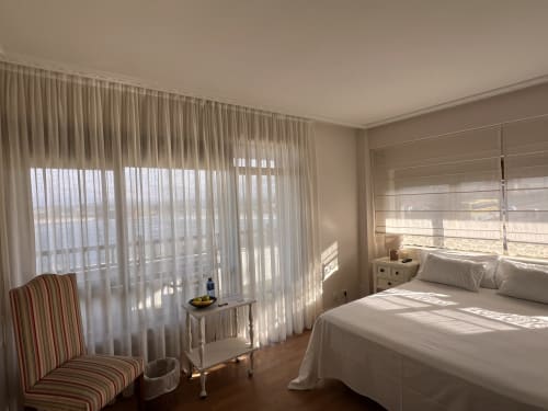 Habitación de hotel luminosa en Playa de Los Locos con vista al mar, cama blanca, silla rayada y cortinas translúcidas que filtran la luz costera.