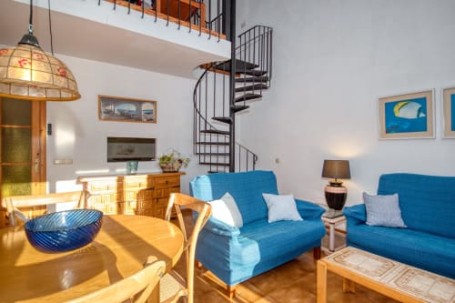Bright apartment living room in Calella with blue sofas, wooden furniture, spiral staircase, and city views at Finques Garcia