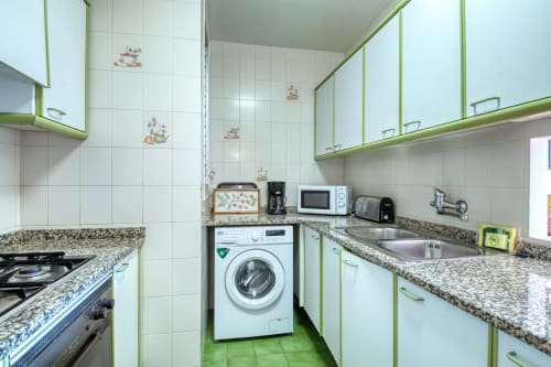 Modern kitchen in Calella apartment with green cabinetry, granite countertops, washing machine, and white tile walls