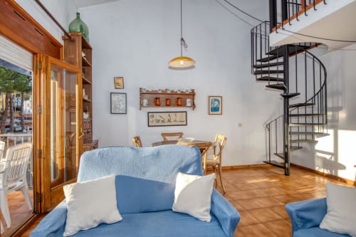 Bright apartment living room in Palafrugell with blue sofa, wooden furniture, spiral staircase, and terracotta flooring