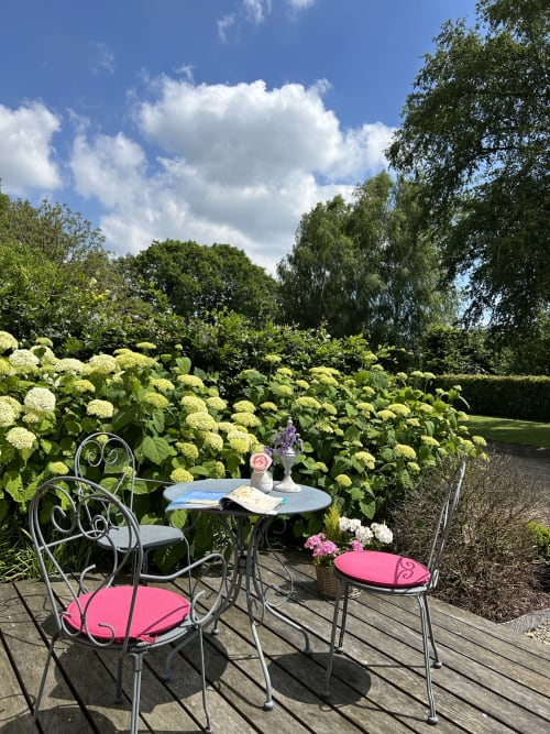 Charmant coin détente du Clos Mahiel avec chaises ornementales, table fer forgée dans le jardin parmi les fleurs bleues et les hortensias luxuriants sous un ciel bleu