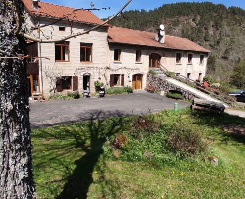 Maison en pierre Au Bord de la Dunière à Sainte-Sigolène avec vue sur la rivière, forêts en arrière-plan et cour d'entrée.
