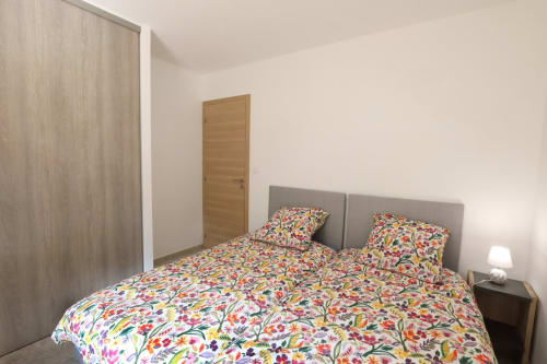 Chambre lumineuse de la maison Au Bord de la Dunière avec parure fleurie colorée, tête de lit grise, portes en bois et lampe moderne