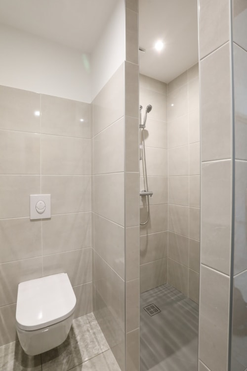Salle de bain moderne avec toilettes blanches, douche à l'italienne avec pomme de douche amovible, carrelage beige et éclairage encastré à Au Bord de