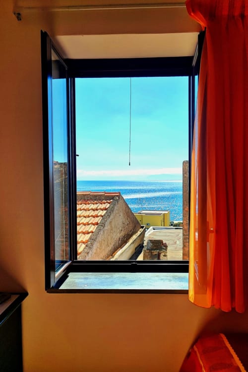 Finestra aperta con vista sul mare dalla camera dell'Hotel Principe di Rinella a Rinella, Italia, con affaccio su tetti in terracotta e blu