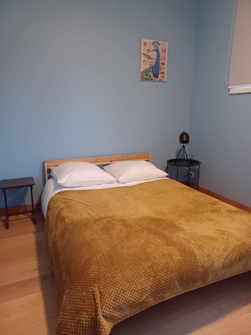 Chambre de l'Hôtel Accolade à Châteaudun avec lit en bois, oreillers blancs, plaid doré et décoration murale paon
