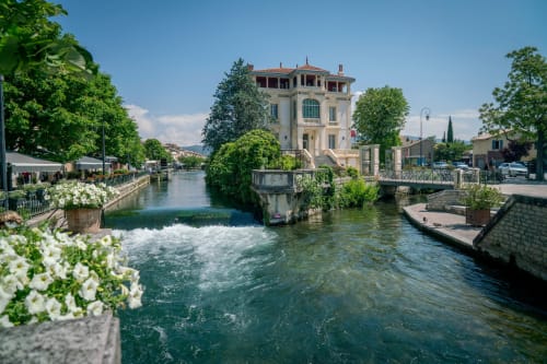Charmante villa au bord de l'eau à L'Isle-sur-la-Sorgue avec rivière turquoise, fleurs blanches et architecture méditerranéenne.