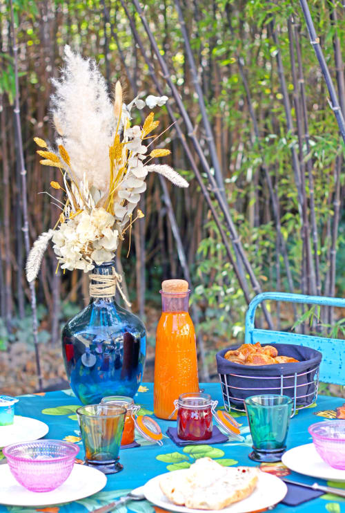 Table dressée en terrasse au Cabanon du Clos avec nappe bleue, composition de fleurs séchées, boissons et vue sur la forêt en Île