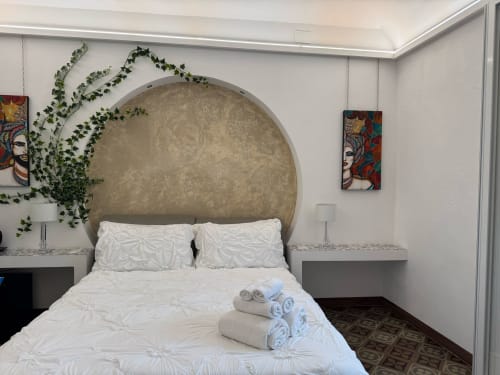 Quarto moderno na pousada I MORI Catania com grande cabeceira arqueada, roupa de cama branca, plantas suspensas e arte de parede colorida