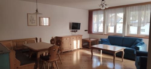 Wohnbereich des Udai Apartments im Residence Rodolon in Mazzin mit Holzesstisch, blauem Sofa, TV und Bergblick