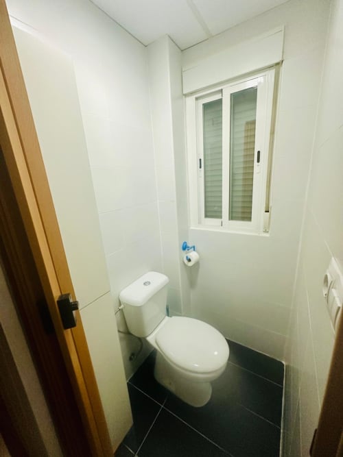 Baño moderno en apartamento de Mérida con paredes de azulejos blancos, inodoro, ventana con persianas y piso de azulejos negros