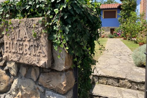 Entrada de Casa La Huerta con letrero de piedra, enredaderas de hiedra y sendero del jardín en Cangas de Onís