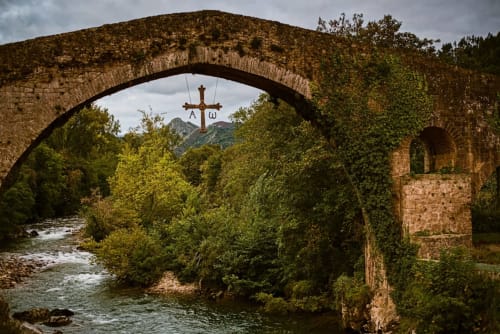 Histórico puente de arco de piedra sobre río con cruz y montaña, Cangas de Onís