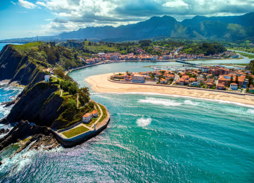 Vista aérea de Cangas de Onís con playa dorada, aguas turquesas, puente histórico y montañas Picos de Europa