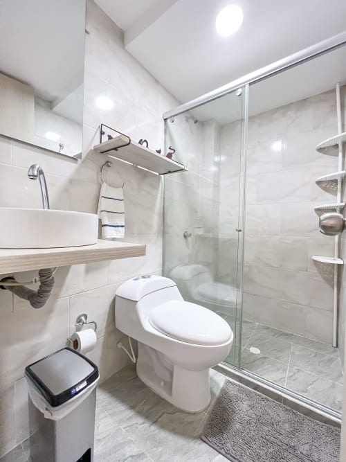 Baño moderno con azulejos blancos, cabina de ducha de cristal, inodoro y lavamanos redondo con tocador de madera en el hotel Olamar Living