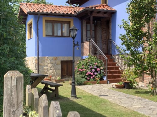 Patio de Casa La Huerta en Cangas de Onís con casa azul, mesa de picnic, flores rosas y camino de piedra