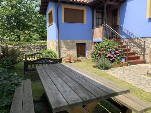 Patio jardín de Casa La Huerta con mesa de picnic de madera, edificio azul, flores rosas y escaleras de piedra en Cangas de Onís
