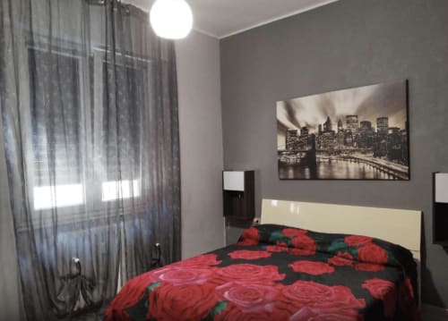 Modern bedroom with red rose bedding, cream headboard, gray walls, city skyline artwork, and sheer curtains at Navigli