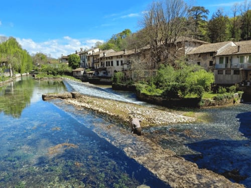 "Scorcio del borgo storico di Polcenigo con il torrente Gorgazzo che crea piccole cascate tra gli edifici antichi, alberi spogli e il ponte pedonale sulla sinistra."