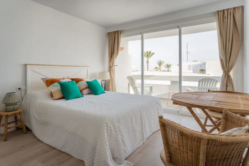 Luminoso dormitorio de estudio en Formentera Especial con balcón con vistas al mar, cama blanca, cojines coloridos y zona de estar de mimbre