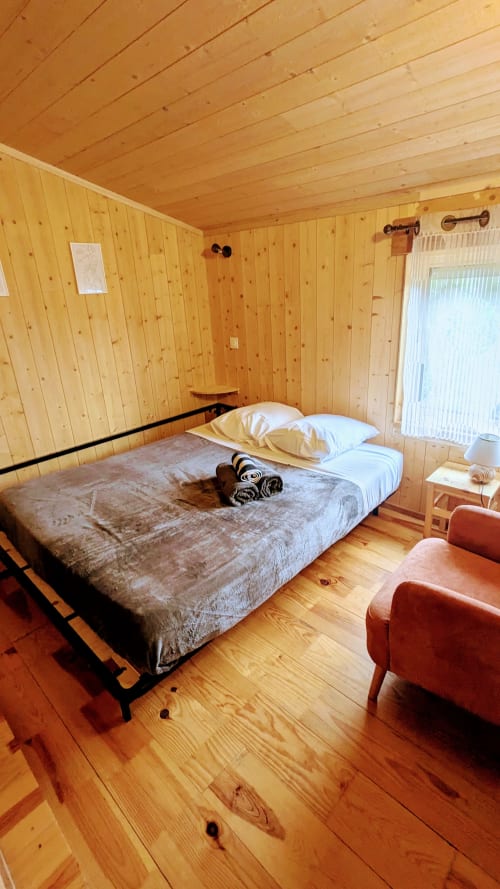 Cozy wooden cabin bedroom at Cabanes du Belvédère with bed, pillows, red chair, and pine interior