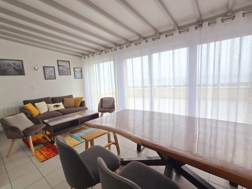 Modern open-plan living room with sea view, brown sofa, dining table, and sheer curtains at Relais des Îles cottage,