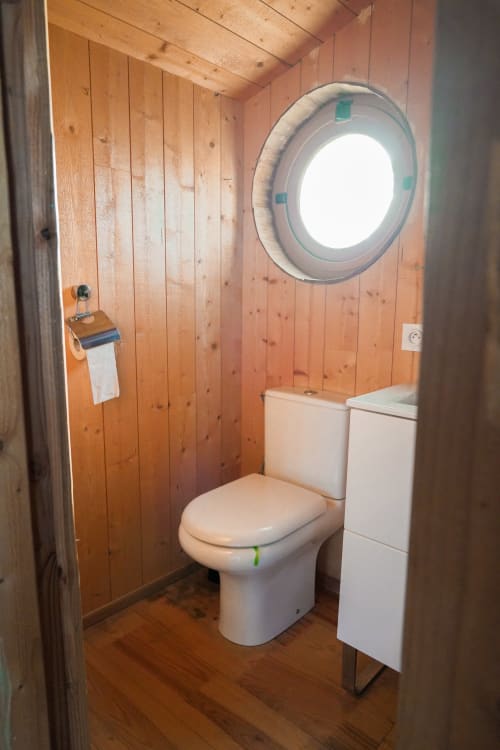 Modern bathroom in wooden cabin with white toilet, sink, and circular porthole window at Cabanes du Belvédère