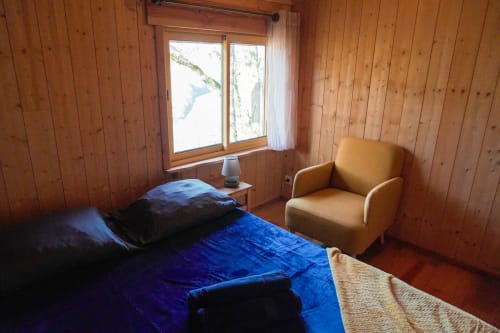 Wooden cabin bedroom with blue bedding, armchair, and lake view window at Cabanes du Belvédère, Serrières-sur-Ain