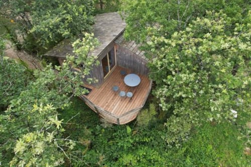Aerial view of wooden treehouse cabin with deck and hot tub surrounded by green forest at Cabanes du Belvédère,
