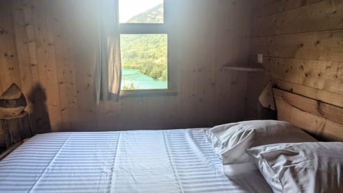 Rustic wooden bedroom with striped bedding overlooking turquoise lake and forested mountains through large window at Cabanes