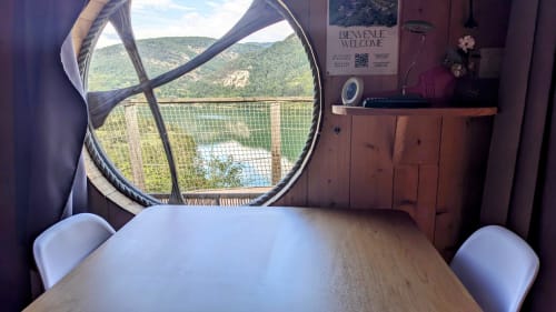 Vista dal tavolo da pranzo attraverso la finestra circolare che si affaccia sul lago e sulle montagne boscose alle Cabanes du Belvédère