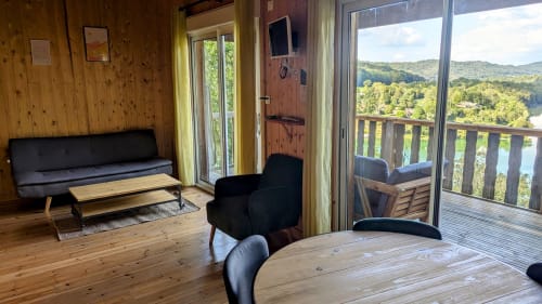 Interno di lodge moderno con pareti in legno, zona soggiorno, tavolo da pranzo e viste panoramiche della valle del fiume Ain e foresta