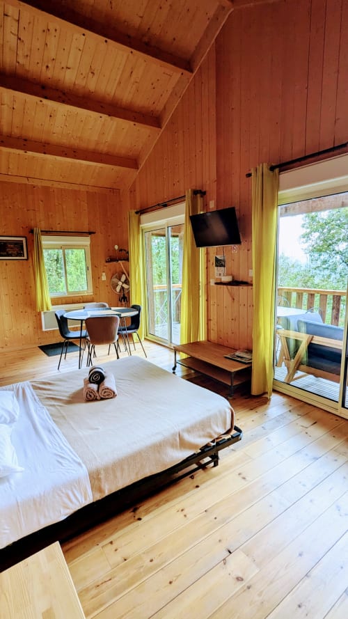 Camera da letto in lodge in legno con letto bianco, tavolo da pranzo, TV a schermo piatto e porte in vetro che si affacciano sul lago a Serrières-sur-Ain
