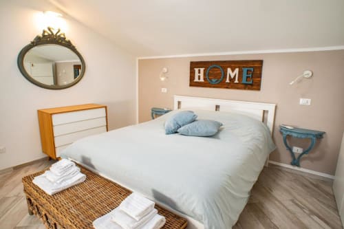 Camera moderna di Ap Appartamenti Bologna con letto matrimoniale, cuscini blu, cassettone in legno, specchio decorativo e cartello HOME a parete